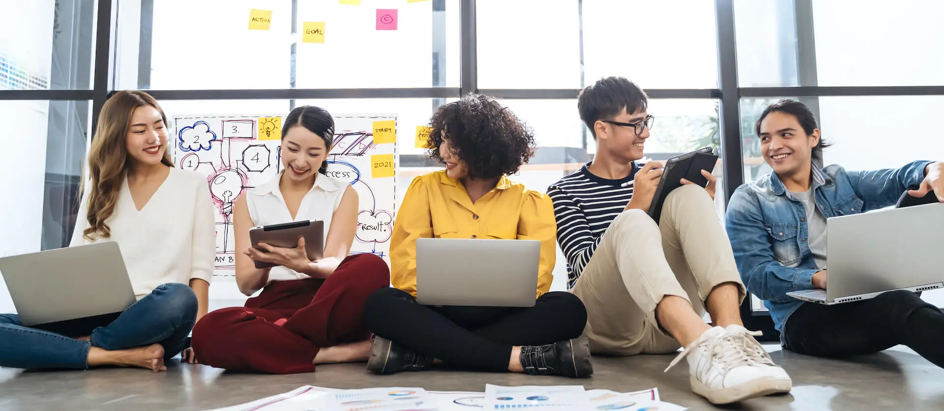 Group of young adults sitting on the floor, collaborating with laptops and tablets in a casual setting, with sticky notes and a whiteboard with diagrams in the background.